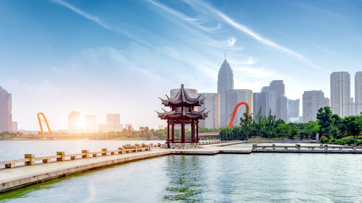 Traditional Pavilion and Modern Skyline in China A scenic view of a traditional Chinese pavilion on a lake walkway, with a modern city skyline and a distinctive orange bridge in the background under a bright sky.